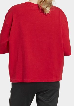 Vaaleahiuksinen henkilö, joka käyttää löysää punaista lyhythihaista paitaa ja mustia housuja valkoisilla raidoilla, kuvattuna takaa.