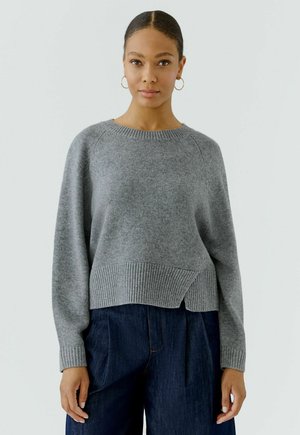 Vrouw met donker haar in een lage knot draagt gouden oorringen, een grijze gebreide trui met geribde zoom en donkerblauwe high-waist broek.