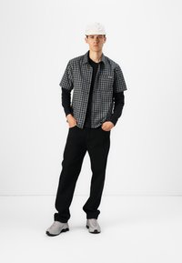 Short-sleeve black and white checkered shirt over a long-sleeve black top, paired with black pants and gray sneakers. White cap completes the outfit.