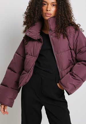 Giacca imbottita color bordeaux con colletto alto, bottoni a scatto e vestibilità ampia, abbinata a una maglietta nera e pantaloni neri su misura.