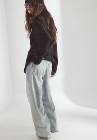 Svart långärmad skjorta med knapppdetaljer som matchas med ljusblå vida jeans. Modellen är accessoariserad med stora mörka solglasögon.