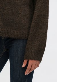 Bruine gebreide trui met een gestructureerde afwerking en geribbelde manchetten, gecombineerd met donkerblauwe spijkerbroek. Focus op de details van de mouwen en de gezellige uitstraling van de stof.