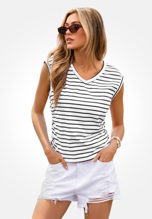 Kobieta w okularach przeciwsłonecznych w oprawkach w kolorze żółwia, w białej i czarnej bluzce bez rękawów w paski oraz w poszarpanych białych szortach jeansowych, z rękami w kieszeniach.