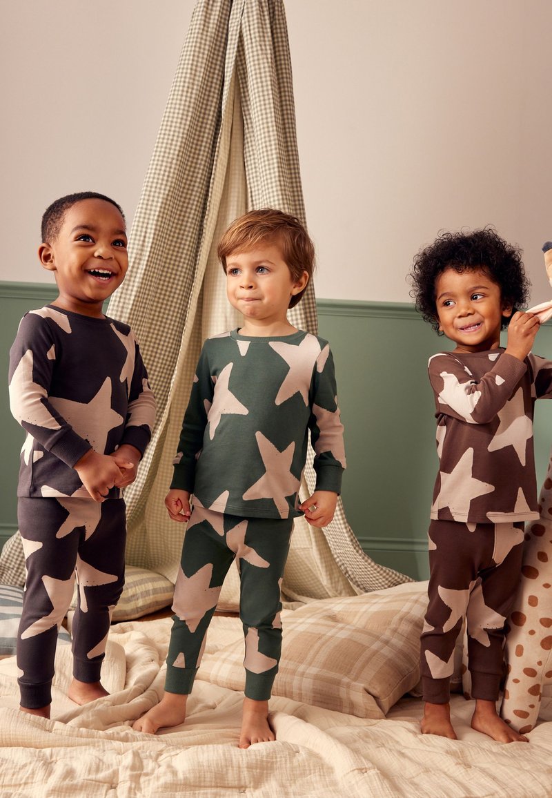Trois tout-petits en pyjamas à motif étoilé sont pieds nus sur une couverture douce devant un tipi à carreaux beige et vert à l’intérieur.