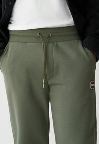 Pantaloni de sudură de culoare verde măslin, dintr-un material texturat, cu un brâu cu șnur, buzunare laterale și un patch circular cu logo pe coapsă.