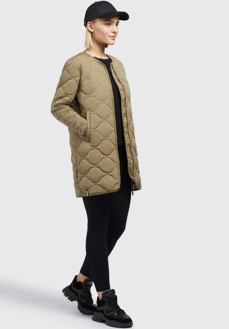 Gepolsterter olivgrüner Mantel mit Rautenmuster, Reißverschluss, seitlichen Taschen sowie kontrastierenden schwarzen Leggings und Sneakers.