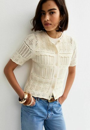 Joven mujer con cárdigan corto de punto color crema con botones, jeans azules, grandes pendientes de aro y pulseras de materiales mixtos, de pie frente a una pared blanca.