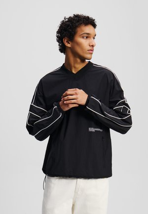 Sort letvægtsjakke med V-hals, der har hvide accentstriber på ærmerne og en sidelukning med lynlås. "KARL LAGERFELD JEANS" logoet er synligt.