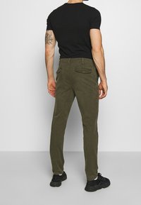 Olive green chinos in cotton twill with a straight fit, featuring two back pockets with button closures, paired with a black T-shirt and black sneakers.