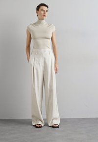 Μπεζ εφαρμοστό ψηλόμεσο μπουφάν σε συνδυασμό με κρεμ παντελόνι πλατύ στους γοφούς. Λείο ύφασμα, πτυχώσεις στο μπροστινό μέρος και μαύρες σαγιονάρες.