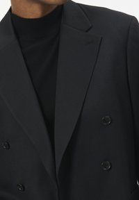 Juodas dvigubos sagtys švarkas su struktūruotais atlapais, turintis subtilią raštuotą tekstūrą ir du juodus mygtukus. Dėvimas ant juodo aukšto kaklo marškinėlių.