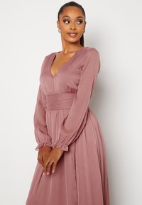 Šaty s dlouhým rukávem z jemné růžové látky, s hlubokým výstřihem do V a přiléhavým pasem, s volnou sukní a nařasenými manžetami rukávů.