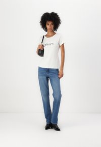 Weißes Kurzarm-T-Shirt mit metallic Aufdruck, kombiniert mit hellblauen geraden Jeans und schwarzen spitzen High Heels, dazu eine kleine schwarze Tasche.