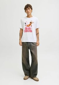 Witte T-shirt met een kleurrijke print van een roze flamingo en een rendier met de tekst "VROLIJK KERSTFEEST". Gecombineerd met vervaagde grijze spijkerbroeken en beige schoenen.
