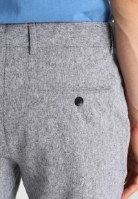 Šedé tkané kalhoty s texturovaným povrchem, které mají zadní kapsu uzavřenou černým knoflíkem. Tkanina má jemný vzor.