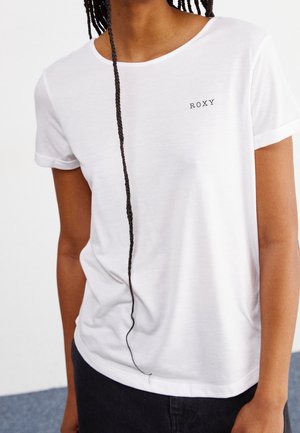 Osoba ubrana w biały T-shirt z krótkim rękawem z napisem „ROXY” na piersi oraz długim, zaplecionym warkoczem zwisającym w dół.