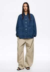 Giacca di denim oversize con grandi tasche frontali, colore azzurro chiaro, chiusura a bottoni e vestibilità comoda, abbinata a pantaloni beige a gamba larga.