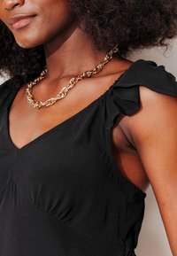 Robe noire avec un décolleté en V, des manches courtes à volants et des détails froncés au niveau de la poitrine. Collier chaîne dorée apportant un accent contrastant.