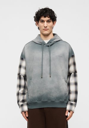 Młody mężczyzna z kręconymi włosami nosi niebiesko-szarą bluzę z kapturem warstwowo na czarno-białej kraciastej koszuli i brązowych spodniach, stojąc na jednolitym tle.