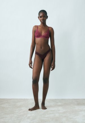 Mujer de pie descalza sobre una alfombra, vistiendo un conjunto a juego de sujetador y panties de encaje burdeos, frente a un fondo liso gris claro.