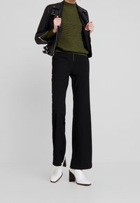 Pantaloni neri a gamba larga, maglione verde a maglia testurizzata, giacca di pelle nera da motociclista e stivaletti bianchi con tacco completano l'outfit.