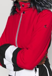 Persona que lleva una chaqueta de invierno roja, negra y blanca con capucha forrada de piel y cierre frontale, con los brazos cruzados sobre el pecho.