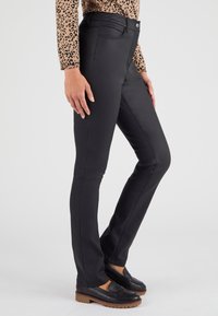 Jeans elásticos negros con cintura alta, que presentan costuras visibles, bolsillos delanteros y un cierre de botón, combinados con mocasines negros.