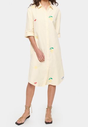 Kobieta ubrana w jasnżółtą, sięgającą kolan sukienkę koszulową zapinaną na guziki, z nadrukiem cytryn i słońca oraz rękawami do łokcia, zestawioną z beżowymi sandałami na paski.