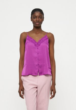 Kvinde iført en lilla ærmeløs bluse med blonder og knapper, kombineret med lyserøde bukser, stående mod en hvid baggrund.