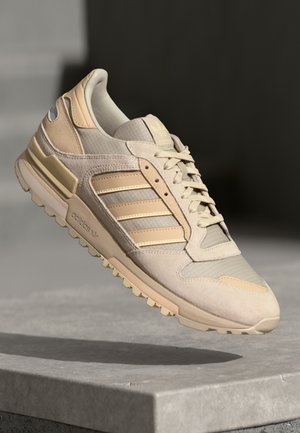 Baskets Adidas beige et crème avec trois bandes et semelle texturée, suspendues au-dessus d'une surface en béton gris clair.