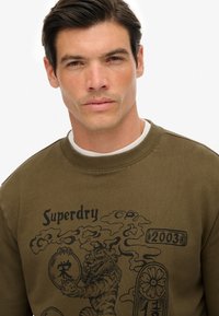 Felpa verde oliva con un audace design grafico nero, che include una tigre e il testo "Superdry 2003". Collo rotondo e bordi a coste.