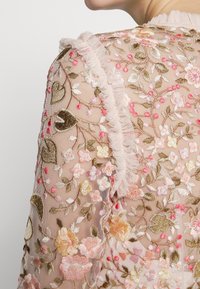 Tissu transparent brodé de motifs floraux et de cœurs en rose, or et vert, avec un galon froncé le long de l'encolure.