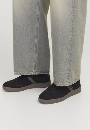 Zapatillas de ante negras con suela de goma texturizada. Presentan un diseño de rayas grises y cordones en la parte delantera. Combinadas con unos vaqueros de mezclilla azul claro.