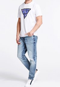 Camiseta blanca con un diseño gráfico triangular morado y texto "GUESS", combinada con jeans claros desgastados y zapatillas blancas.
