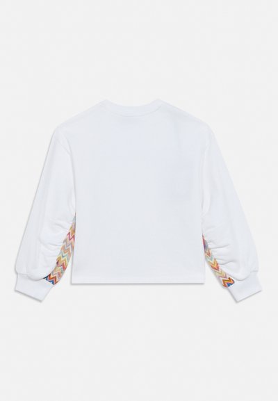 Λευκό πουκάμισο με μακριά μανίκια και πολύχρωμα ζιγκ-ζαγκ σχέδια κατά μήκος των εσωτερικών πλευρών και των δύο μανικιών σε λευκό φόντο.