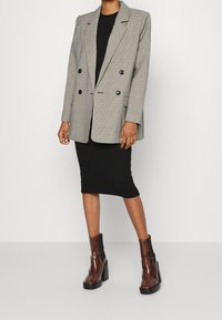 Grau karierter Doppelknopf-Blazer mit spitzen Revers, kombiniert mit einem schwarzen gerippten Kleid und braunen, hochhackigen Stiefeletten.