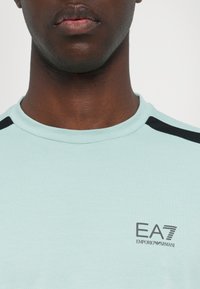 Svetlo modra kratka majica s kratkimi rokavi, okroglim ovratnikom in črnimi poudarki na ramenskih delih, z logotipom EA7 Emporio Armani na sprednji strani.