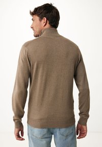Maglione marrone lavorato a maglia con colletto alto, polsini e orlo a coste. Il materiale sembra morbido con una texture liscia. Indossato con jeans azzurri chiari.
