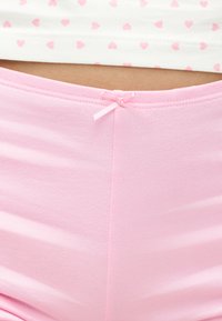 Gros plan sur un pantalon de pyjama en coton rose avec un petit nœud à la taille, associé à un haut blanc avec des motifs de cœurs roses.