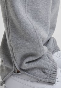 Hallid dressipüksid elastsete varrukatega ja külgnooltega, kantud valgete pitsidega spordijalatsite kohal heledal taustal.