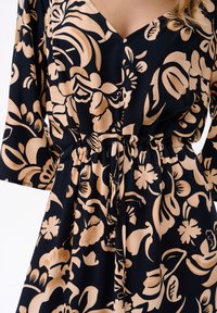 Granatowa sukienka z beżowym kwiatowym wzorem, dekoltem w serek, rękawami o długości 3/4 oraz paskiem w talii do regulacji.