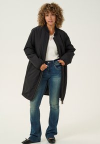 Schwarzer, langer Daunenmantel mit hohem Kragen und gerippten Bündchen. Reißverschluss und Seitentaschen. Kombiniert mit hellblauen Schlagjeans und flachen Schuhen.