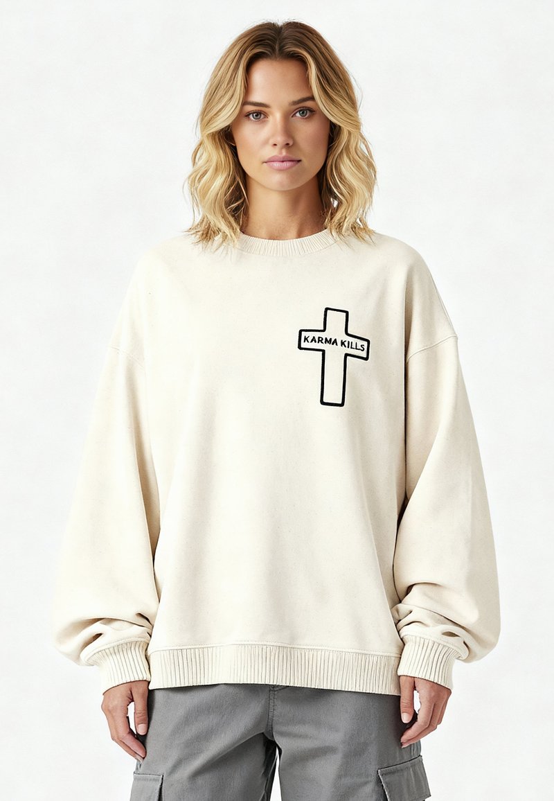 Jeune femme aux cheveux blonds mi-longs portant un sweat-shirt ample blanc cassé avec une croix noire et le texte « KARMA KILLS ».