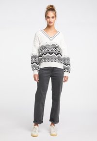 Weißer Cable-Knit-Pullover mit schwarzen geometrischen Mustern. V-Ausschnitt, lockerer Schnitt und Fransenverzierungen am Saum. Kombiniert mit grauen Jeans und weißen Sneakers.