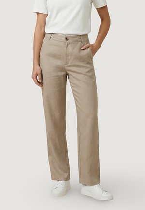 Persona che indossa pantaloni beige a gamba dritta, maglietta bianca a maniche corte con scollo tondo e sneakers bianche con lacci, in piedi con una mano in tasca.