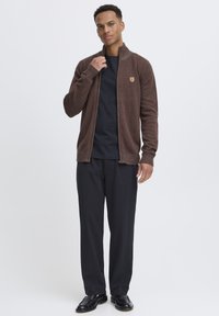 Braune, Reißverschluss-Kardigan mit geripptem Ausschnitt und Bündchen, getragen über einem schwarzen Hemd, kombiniert mit schwarzen Hosen und Schuhen. Einfaches Design, keine Muster.
