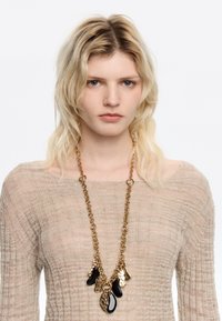 Collier en chaîne dorée comportant plusieurs pendentifs abstraits noirs et dorés. La chaîne a une finition texturée et les pendentifs varient en forme et en taille.
