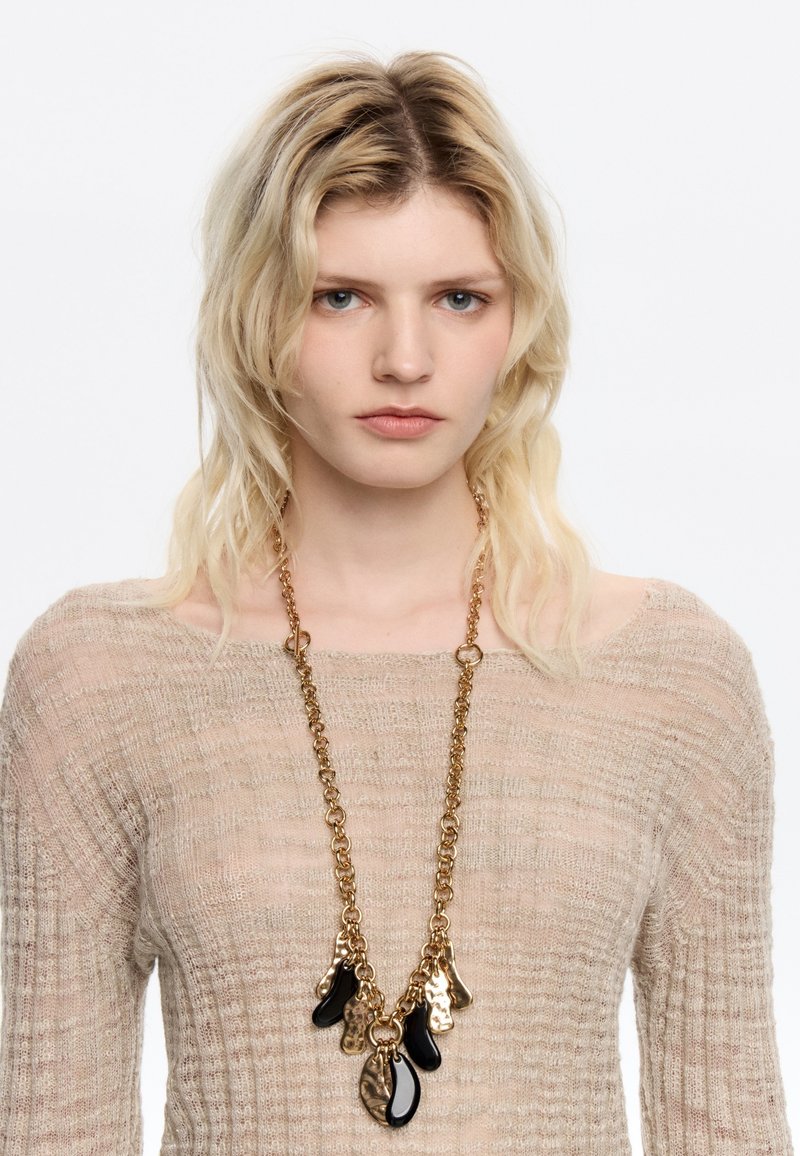 Collier en chaîne dorée comportant plusieurs pendentifs abstraits noirs et dorés. La chaîne a une finition texturée et les pendentifs varient en forme et en taille.