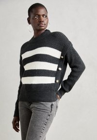 Svarte og hvite stripe strikkegenser med høy hals, ribbestrikket tekstur og knappdetaljer på siden. Brukt sammen med grå jeans.