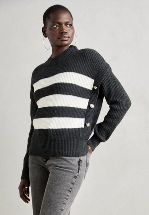 Pull en tricot rayé noir et blanc avec un col montant, texture côtelée et accents de boutons sur le côté. Porté avec un jean gris.